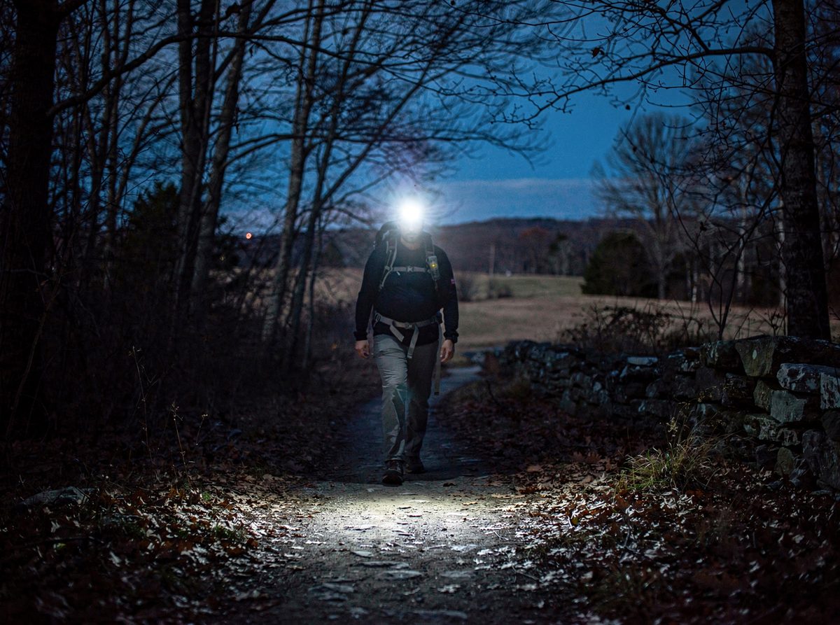 A hiker wearing a headlamp walks a narrow trail at dusk with the beam lighting the tread